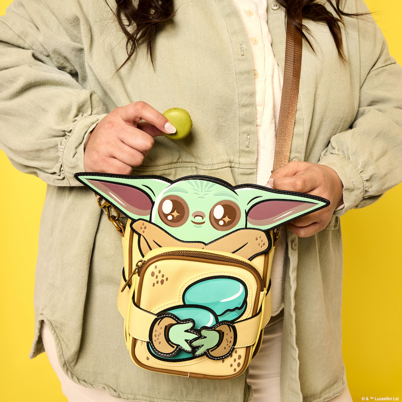 Brunette woman standing against a yellow background holding a green macaron cookie while wearing the Limited Edition Loungefly The Mandalorian Grogu Crossbuddies Bag, with the coin purse showing Grogu holding cookies. 